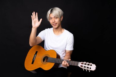 Siyah arka planda gitar çalan Asyalı genç adam mutlu bir ifadeyle selamlıyor.