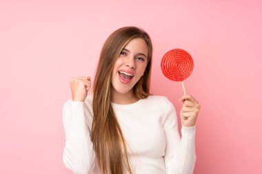 Genç sarışın kız, izole edilmiş pembe arka planda lolipop tutarak zaferi kutluyor.