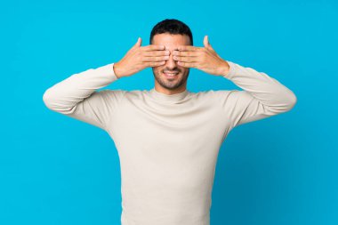 İzole edilmiş mavi arka planı olan genç bir adam gözleri elleriyle kapatıyor.