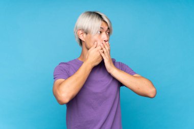 Asyalı genç bir adam izole edilmiş mavi arka planda ağzını kapatıyor ve yana bakıyor.