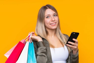 Sarışın, izole edilmiş, sarı arka planı olan genç bir kadın alışveriş torbaları tutuyor ve bir arkadaşına cep telefonuyla bir mesaj yazıyor.