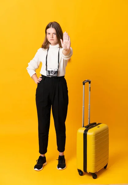 Full body of traveler teenager girl with suitcase over isolated yellow background making stop gesture with her hand