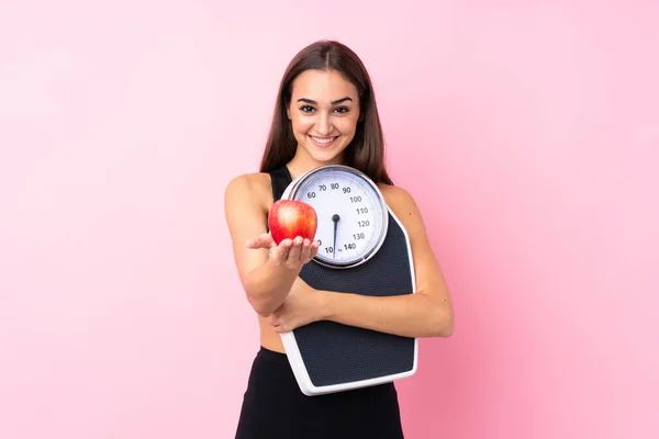 Pretty young girl with weighing machine over isolated pink background holding weighing machine and offering an apple