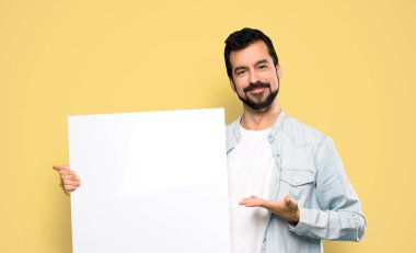 Handsome man with beard holding an empty placard over isolated yellow background