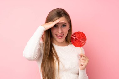 Genç sarışın kız pembe arka planda lolipop tutuyor. Şaşırmış ve şok olmuş bir yüz ifadesi var.