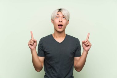 Tecrit edilmiş yeşil arka planı olan genç Asyalı adam şaşırmış ve yukarı bakıyor.