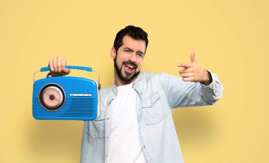 Handsome man with beard holding a radio over isolated yellow background