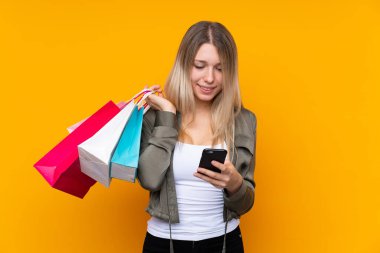 Sarışın, izole edilmiş, sarı arka planı olan genç bir kadın alışveriş torbaları tutuyor ve bir arkadaşına cep telefonuyla bir mesaj yazıyor.