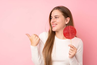 Genç sarışın kız, izole edilmiş pembe arka planda lolipop tutuyor ve bir ürün sunmak için yan tarafı işaret ediyor.