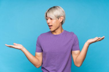 İzole edilmiş mavi arka plan üzerinde genç Asyalı adam iki eliyle kopyalama alanını tutuyor.