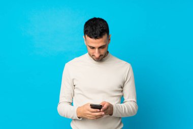 İzole edilmiş mavi arka planda genç bir adam cep telefonuyla mesaj gönderiyor.
