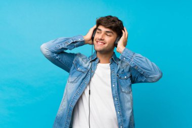 Soyutlanmış mavi arka planda genç yakışıklı adam kulaklıklı cep telefonunu kullanıyor.