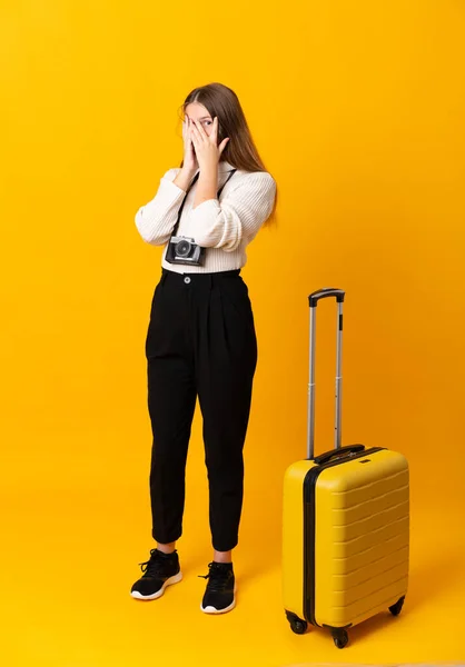 Full body of traveler teenager girl with suitcase over isolated yellow background covering eyes and looking through fingers