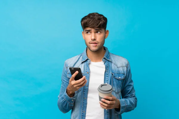 Młody Przystojny Mężczyzna Odizolowanym Niebieskim Tle Trzymając Kawę Zabrania Komórkę — Zdjęcie stockowe
