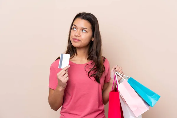 若いですコロンビアの女の子とともにショッピングバッグ上の隔離された背景保持ショッピングバッグとクレジットカードと思考 — ストック写真