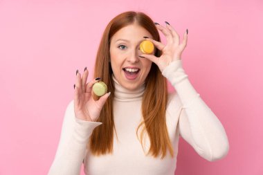 Genç kızıl saçlı, soyutlanmış pembe arka planda, elinde renkli Fransız kurabiyeleri olan bir kadın.