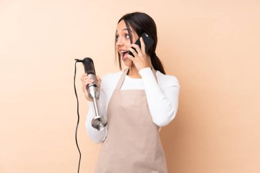 Genç esmer kız el blenderi kullanarak izole bir arkaplanda cep telefonuyla sohbet ediyor.