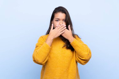 İzole edilmiş mavi arka planı olan genç Kolombiyalı kız elleriyle ağzını kapatıyor.