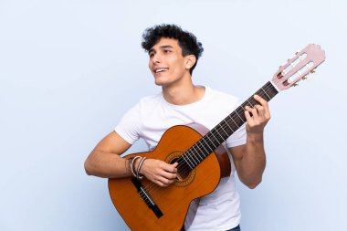 İzole edilmiş mavi arka planda gitar çalan genç Arjantinli adam.