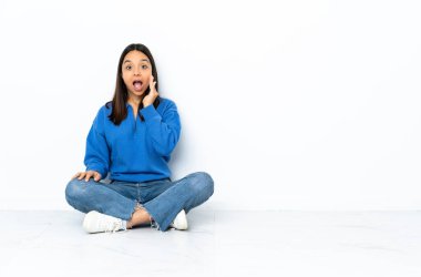 Yerde oturan genç melez kadın beyaz arka planda yalnız ve şaşkın bir yüz ifadesiyle.