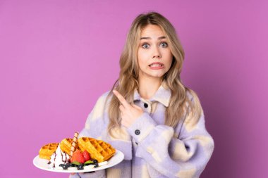 Mor arka planda izole edilmiş waffle tutan genç Rus kız korkmuş ve yan tarafı işaret ediyor.