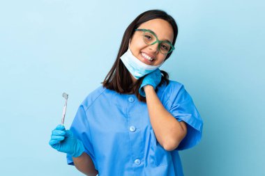 Genç, esmer, melez bir dişçi kadın elinde aletlerle arka planda gülüyor.