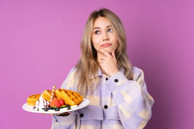 Mor arka planda izole edilmiş waffle tutan genç Rus kız kuşkuları ve kafa karıştırıcı bir ifadeyle