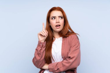 İzole edilmiş mavi arka plandaki kızıl saçlı genç kız bir fikrin parmağı yukarı kaldırdığını düşünüyor.
