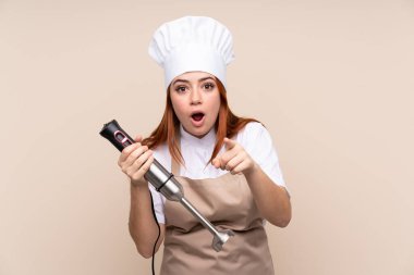 Kızıl saçlı genç kız el blenderi kullanıyor. Tecrit edilmiş arka plan üzerinde.