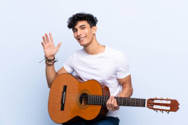 Gitarlı genç Arjantinli adam mavi arka planda mutlu bir ifadeyle selamlıyor.