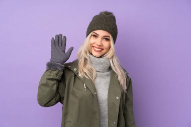 Kış şapkalı genç sarışın kız izole edilmiş mor arka planda mutlu bir ifadeyle selam veriyor.