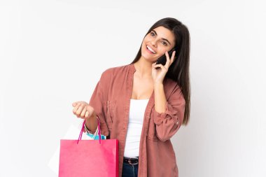 Soyutlanmış beyaz arka planda genç bir kadın alışveriş torbaları tutuyor ve cep telefonuyla bir arkadaşını arıyor.