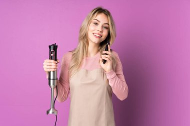 Mor arka planda izole edilmiş el blenderi kullanan genç Rus kız cep telefonuyla biriyle sohbet ediyor.