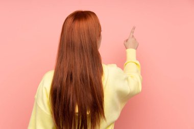 İzole edilmiş pembe arka planı olan kızıl saçlı genç kız işaret parmağıyla işaret ediyor.