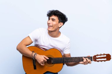 İzole edilmiş mavi arka planda gitar çalan genç Arjantinli adam.