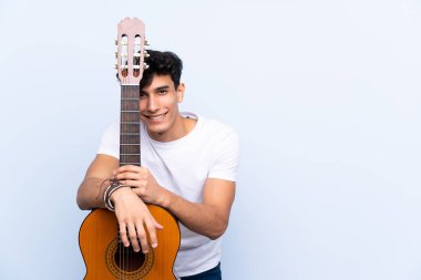 İzole edilmiş mavi arka planda gitar çalan genç Arjantinli adam.