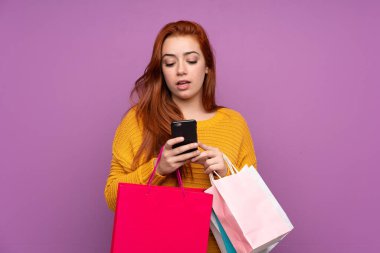 Mor arka planda kalan kızıl saçlı genç kız elinde alışveriş torbalarıyla bir arkadaşına cep telefonuyla bir mesaj yazıyor.
