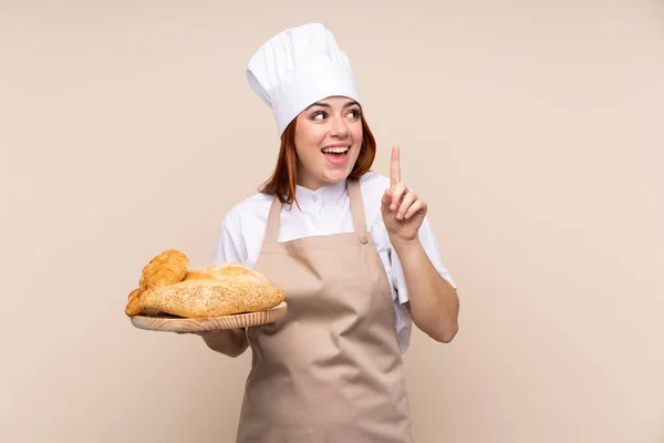 シェフの制服を着た赤毛のティーンエイジャーの女の子 解決策を実現しようとするいくつかのパンとテーブルを保持している女性パン屋指を持ち上げながら — ストック写真