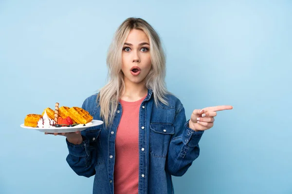 Adolescente Menina Segurando Waffles Sobre Isolado Fundo Azul Surpreso Apontando — Fotografia de Stock