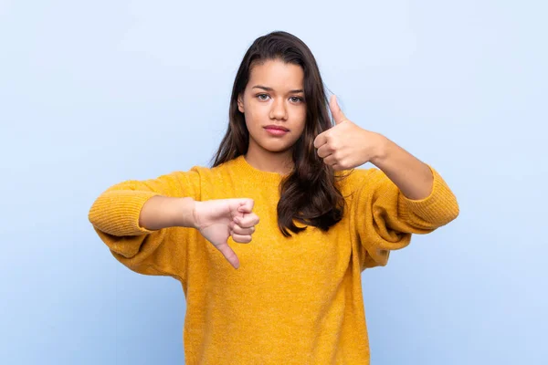 Young Colombian girl with sweater over isolated blue background making good-bad sign. Undecided between yes or not