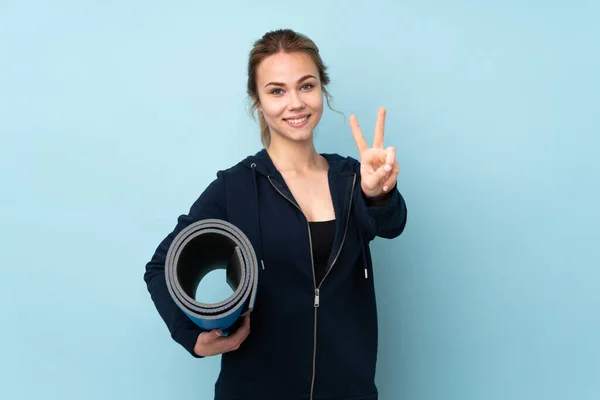 ティーンロシア女の子保持マット隔離上の青背景笑顔とショー勝利サイン — ストック写真