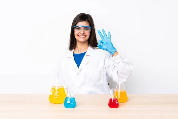 一个年轻的科学女人坐在桌子上 手指显示出一个好的标志 — 图库照片