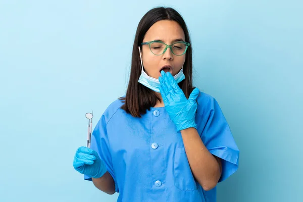 年轻的黑发混血女牙医手拿着工具 在孤立的背景下打呵欠 用手捂住大大的张开的嘴 — 图库照片