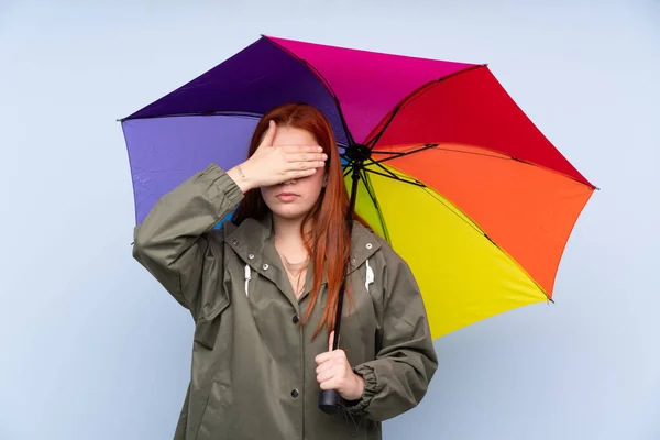 Redhead teenager girl holding an umbrella over isolated blue background covering eyes by hands