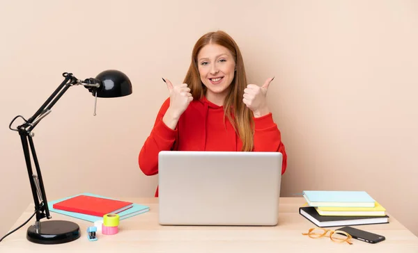 Young student woman in a workplace with a laptop with thumbs up gesture and smiling