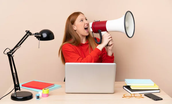 Young student woman in a workplace with a laptop shouting through a megaphone