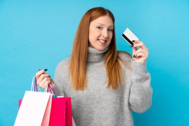İzole edilmiş mavi arka planı olan genç kızıl saçlı kadın elinde alışveriş çantaları ve bir kredi kartı tutuyor.