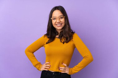 Mor arka planda izole edilmiş, kolları kalçasında ve gülümseyerek poz veren genç melez kadın.