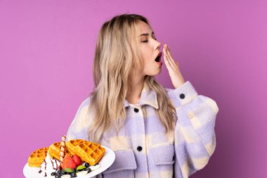 Mor arka planda izole edilmiş waffleları tutan genç Rus kız esniyor ve ağzını eliyle açık tutuyor.