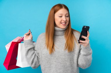 Genç kızıl saçlı kadın, mavi arka planda, elinde alışveriş çantaları ve cep telefonuyla bir arkadaşına mesaj yazıyor.
